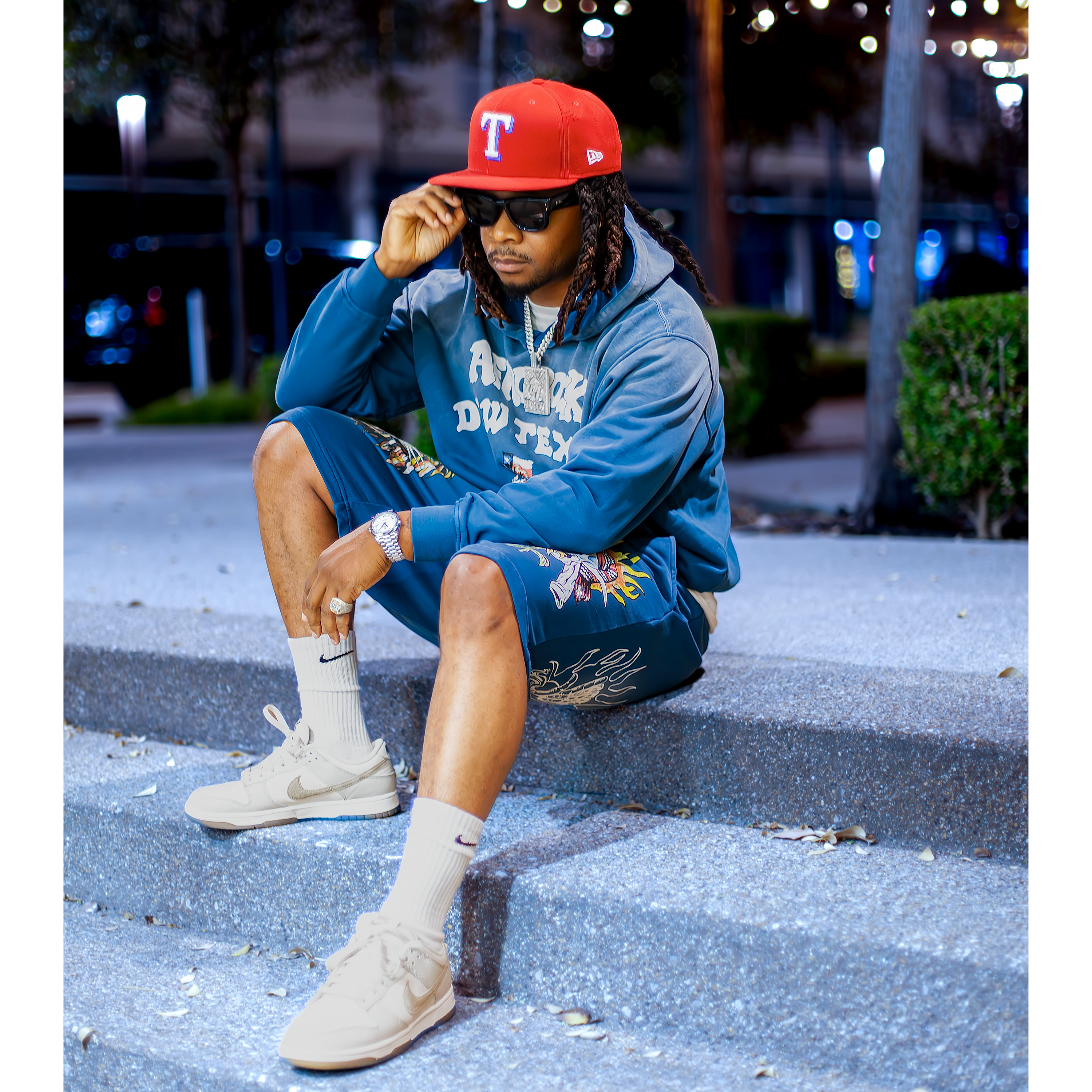 Kal Afrorock wearing the AfroRock DFW TEXAS blue hoodie and shorts with graphic designs and red Rangers hat sitting on a street at night.