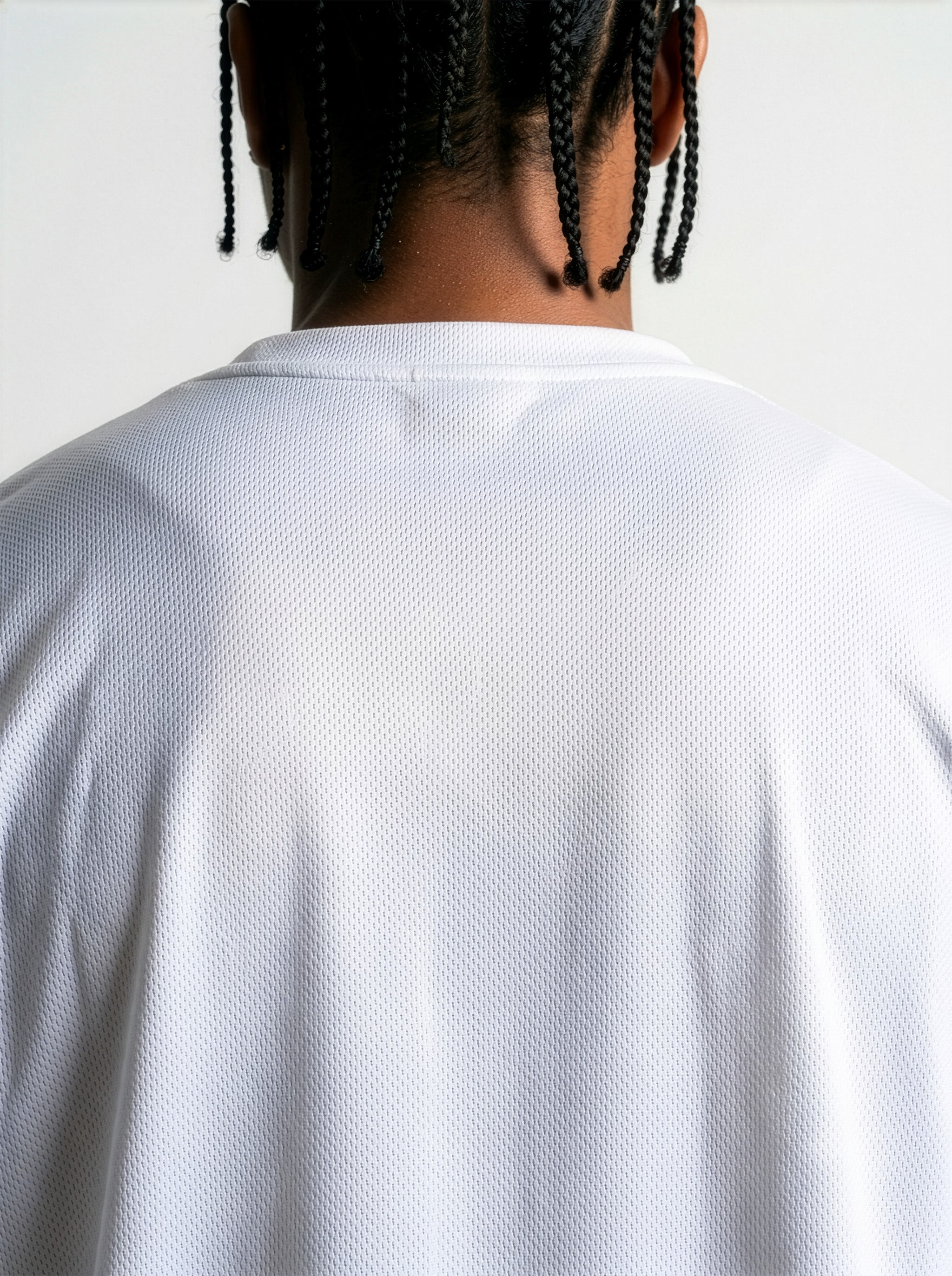 Rear View Commercial studio catalog photography of model Jason featuring sharp box braids, wearing the AfroRock Core Performance T-Shirt in White. Low-angle front-facing view highlighting the minimalist crewneck collar detailing, structured shoulders, and athletic drape of the performance fabric. Clean high-key medium-gray studio background.