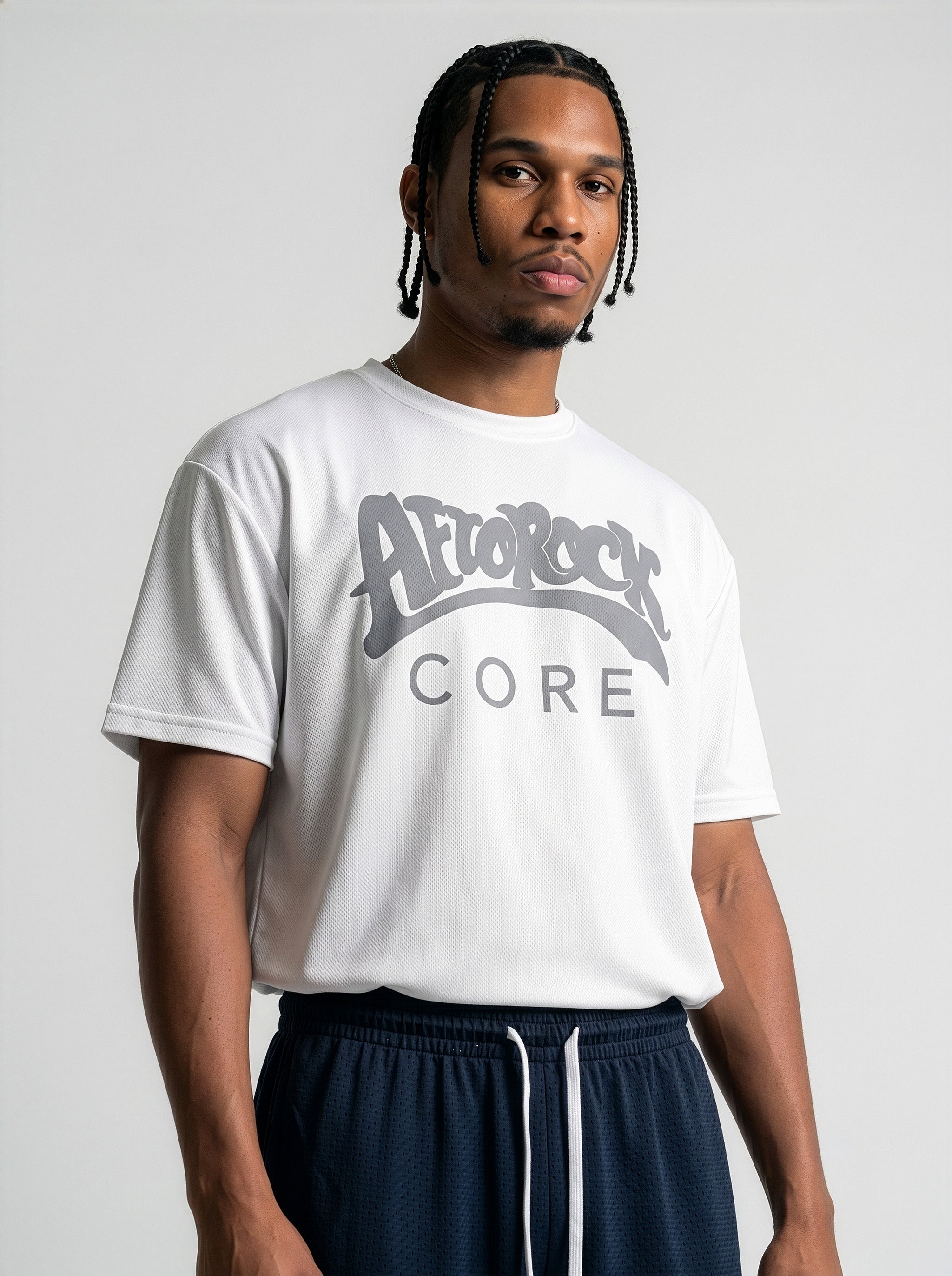 Commercial studio catalog photography of model Jason featuring sharp box braids, wearing the AfroRock Core Performance T-Shirt in White. Low-angle front-facing view highlighting the minimalist crewneck collar detailing, structured shoulders, and athletic drape of the performance fabric. Clean high-key medium-gray studio background.