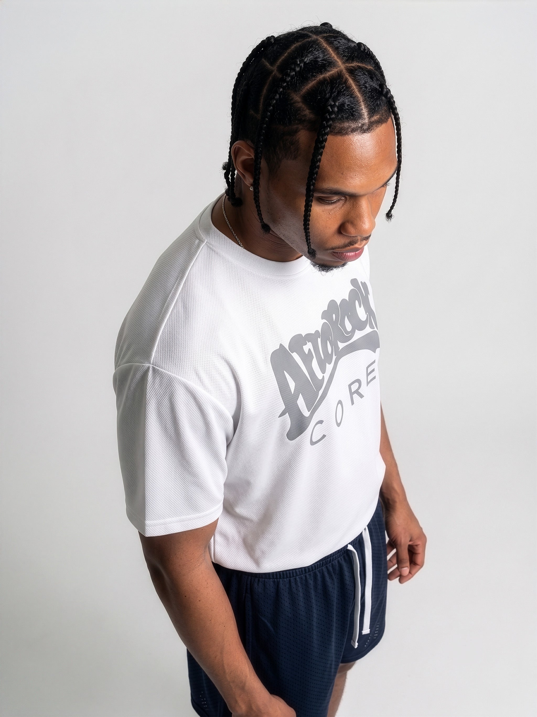 Top View Commercial studio catalog photography of model Jason featuring sharp box braids, wearing the AfroRock Core Performance T-Shirt in White. Low-angle front-facing view highlighting the minimalist crewneck collar detailing, structured shoulders, and athletic drape of the performance fabric. Clean high-key medium-gray studio background.