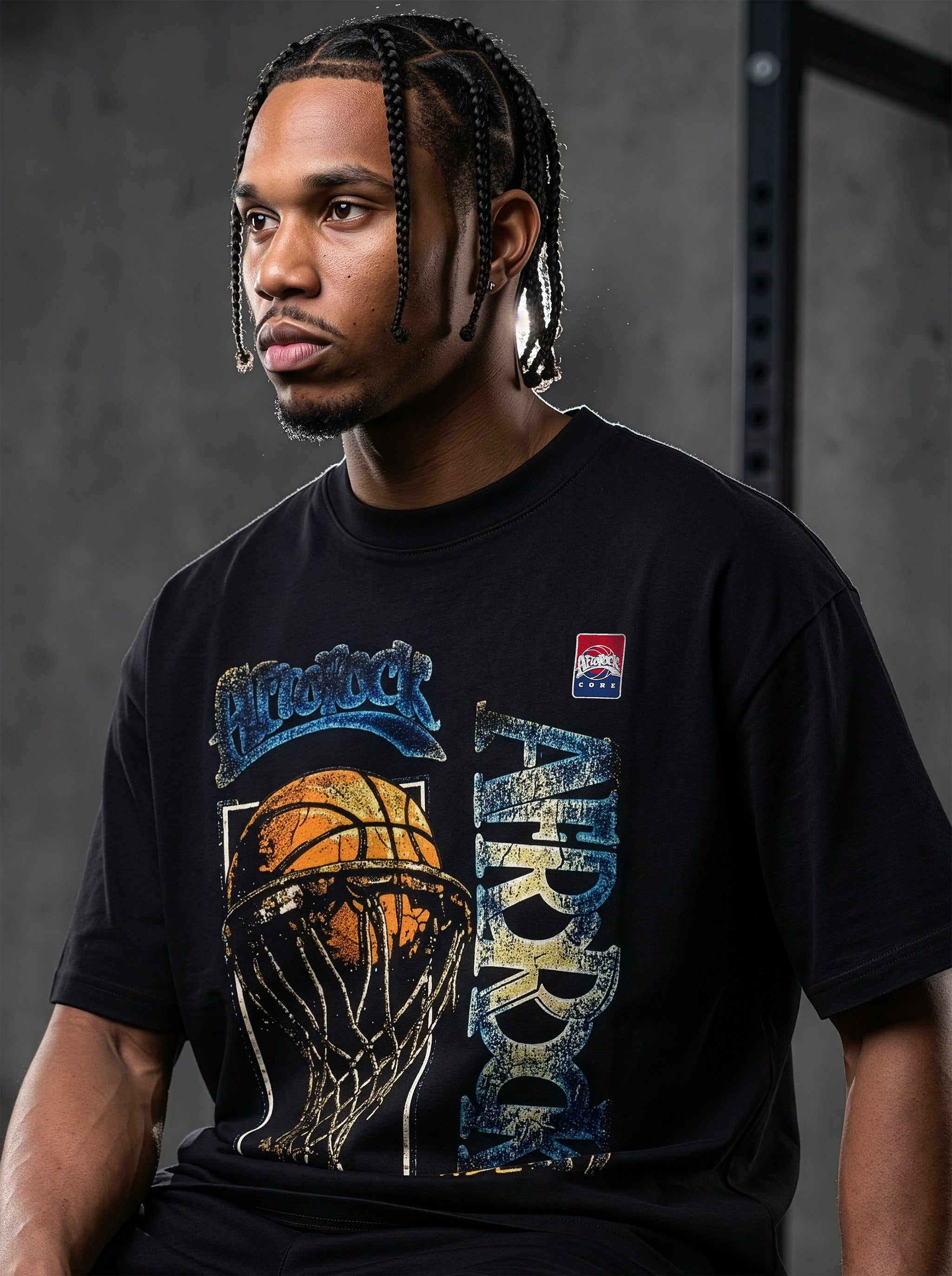 Studio product photography of model Jason in the AfroRock Core streetwear collection. He wears the AfroRock Basketball Vintage Graphic Tee in Pitch Black, an oversized 260gsm cotton shirt with an expansive front graphic of a basketball and hoop, a vintage weather-crackle print finish, and an athletic team badge. Industrial studio lighting highlights a premium sport-heritage lifestyle aesthetic.