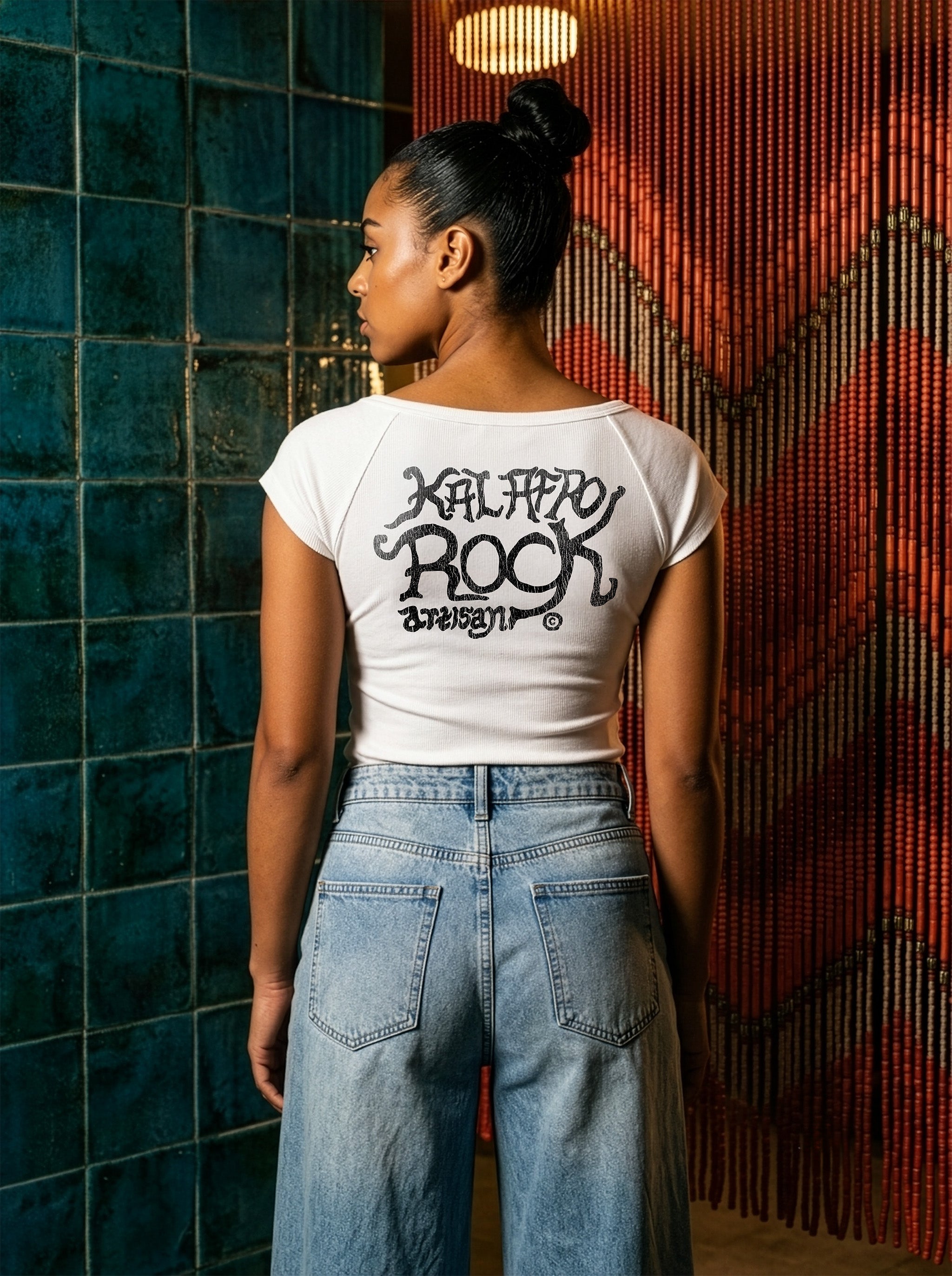 Rear View of Model Kayla in the AfroRock Chief Skull Horns Wide Neck Baby Tee in White. Fine-ribbed bodycon graphic tee featuring an original hand-drawn 1883 West African heritage skull design and artisan back branding by AfroRock.